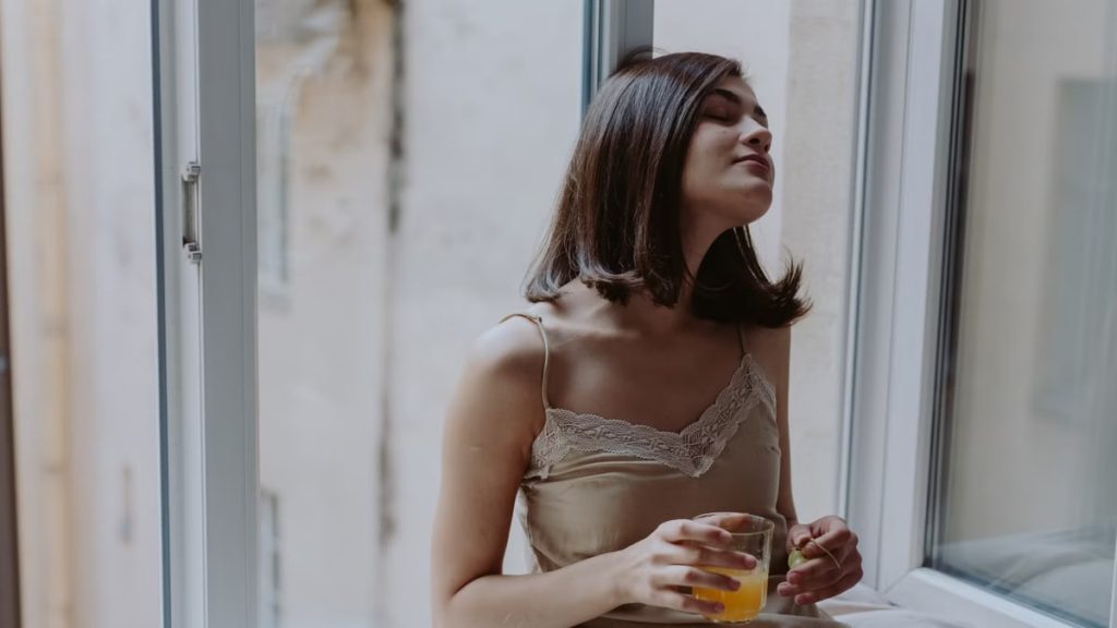 Woman in a spaghetti strap dress savoring orange juice on a window sill, indoors.