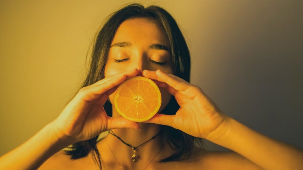 Calm young female with bare shoulders standing with eyes closed against plain wall and covering lips with orange slice
